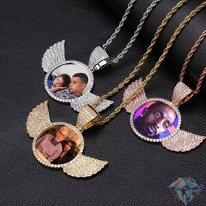 Può includere: Tre collane con ciondoli fotografici personalizzati. Ogni ciondolo presenta una foto circolare circondata da un design ad ali, incastonato con piccole gemme trasparenti. Le catene sono argento, oro e oro rosa.