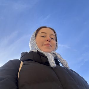 Puede incluir: Una mujer con una chaqueta acolchada negra, un gorro y una bufanda de punto azul claro. El fondo es un cielo azul brillante.