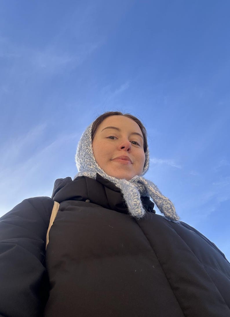 Puede incluir: Una mujer con una chaqueta acolchada negra, un gorro y una bufanda de punto azul claro. El fondo es un cielo azul brillante.