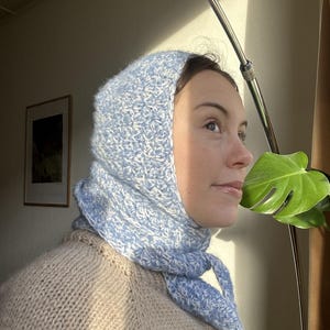 Puede incluir: Una mujer lleva un gorro y una bufanda de ganchillo azul claro y blanco. El gorro est&aacute; tirado sobre las orejas y la bufanda est&aacute; atada alrededor del cuello. Lleva un su&eacute;ter beige.