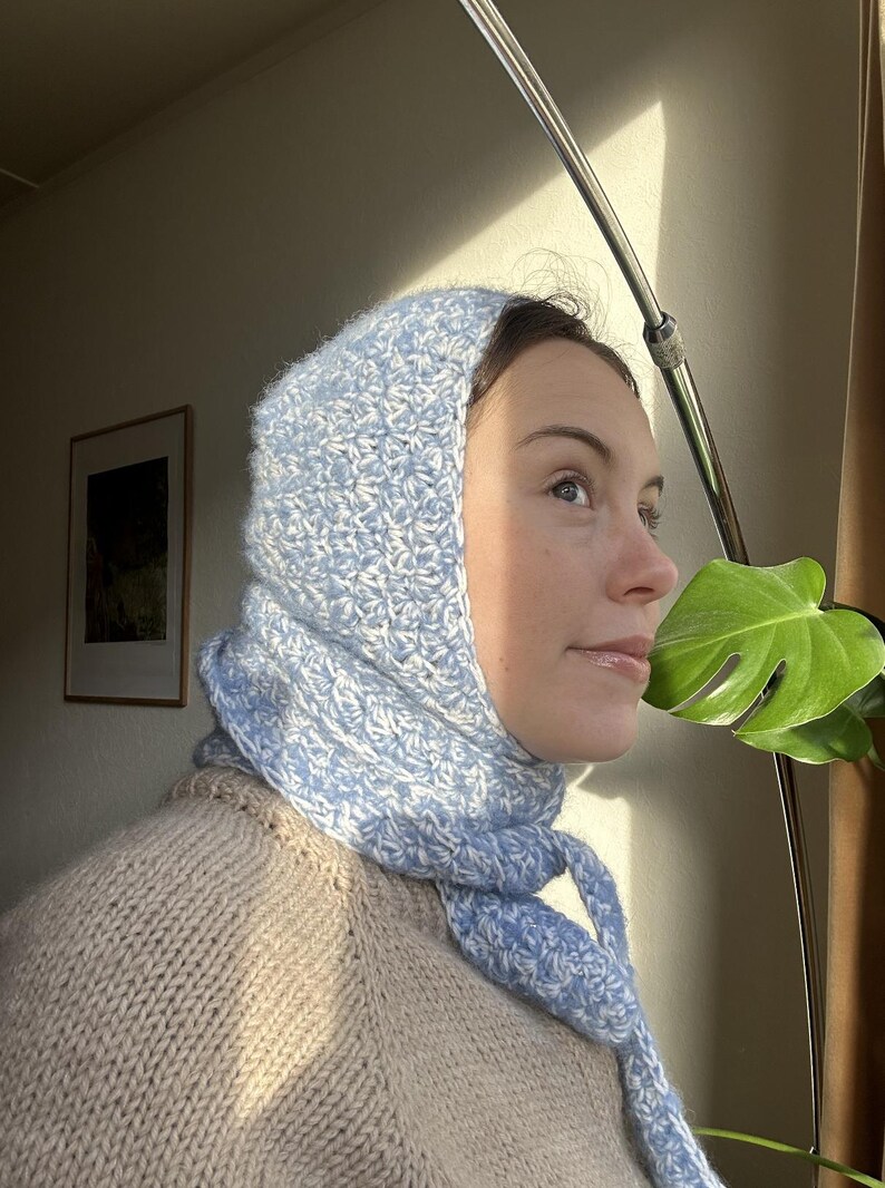Puede incluir: Una mujer lleva un gorro y una bufanda de ganchillo azul claro y blanco. El gorro est&aacute; tirado sobre las orejas y la bufanda est&aacute; atada alrededor del cuello. Lleva un su&eacute;ter beige.