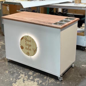 May include: A white and wood-toned mobile bar with a wooden countertop and three stainless steel inserts. The bar features a circular illuminated sign that reads "Your logo here" with a tomato and cocktail glass graphic. The bar is on wheels.