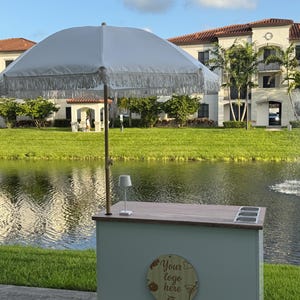 May include: A light blue outdoor bar cart with a white fringed umbrella. The cart has a wooden countertop, three recessed containers, and a small lamp. A wooden sign on the front reads "Your logo here."