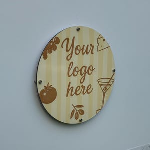 May include: A round wooden sign with a cream and tan striped background. The sign has the words "Your logo here" in a brown script font. It features illustrations of a tomato, grapes, cheese, an olive branch, and a martini.