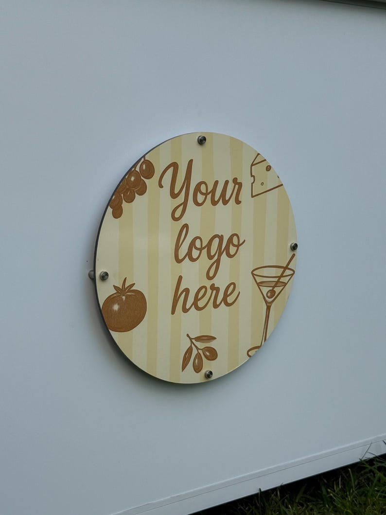 May include: A round wooden sign with a cream and tan striped background. The sign has the words "Your logo here" in a brown script font. It features illustrations of a tomato, grapes, cheese, an olive branch, and a martini.