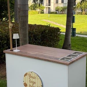 May include: A white outdoor bar with a wooden countertop and three stainless steel inserts. The bar has a white umbrella with fringe and a wooden sign that says "Your logo here". A small lamp sits on the counter.