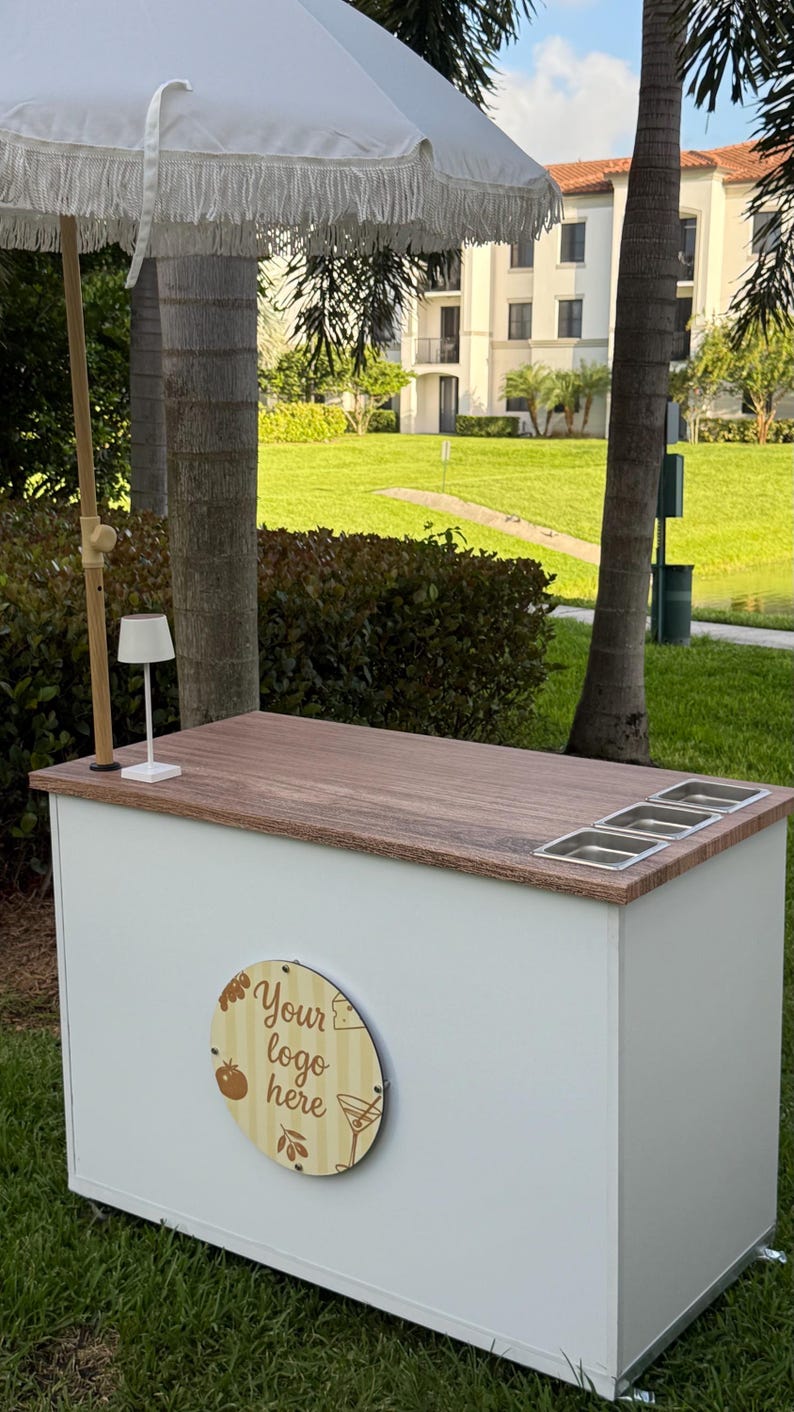 May include: A white outdoor bar with a wooden countertop and three stainless steel inserts. The bar has a white umbrella with fringe and a wooden sign that says "Your logo here". A small lamp sits on the counter.