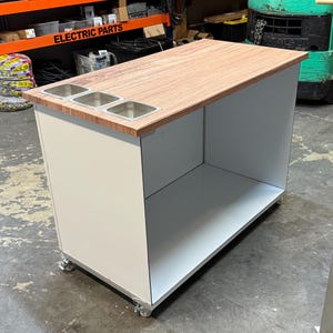 May include: A white and wood-toned rolling cart with three stainless steel containers built into the wood countertop. The cart has an open storage area and is on wheels. The words "ELECTRIC PARTS" are visible on a shelf in the background.