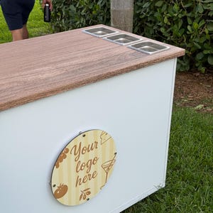 May include: A white and wood-toned outdoor bar with three stainless steel food containers. The bar features a wooden oval sign with the text "Your logo here" and food-related illustrations. The bar is set on a grassy area.