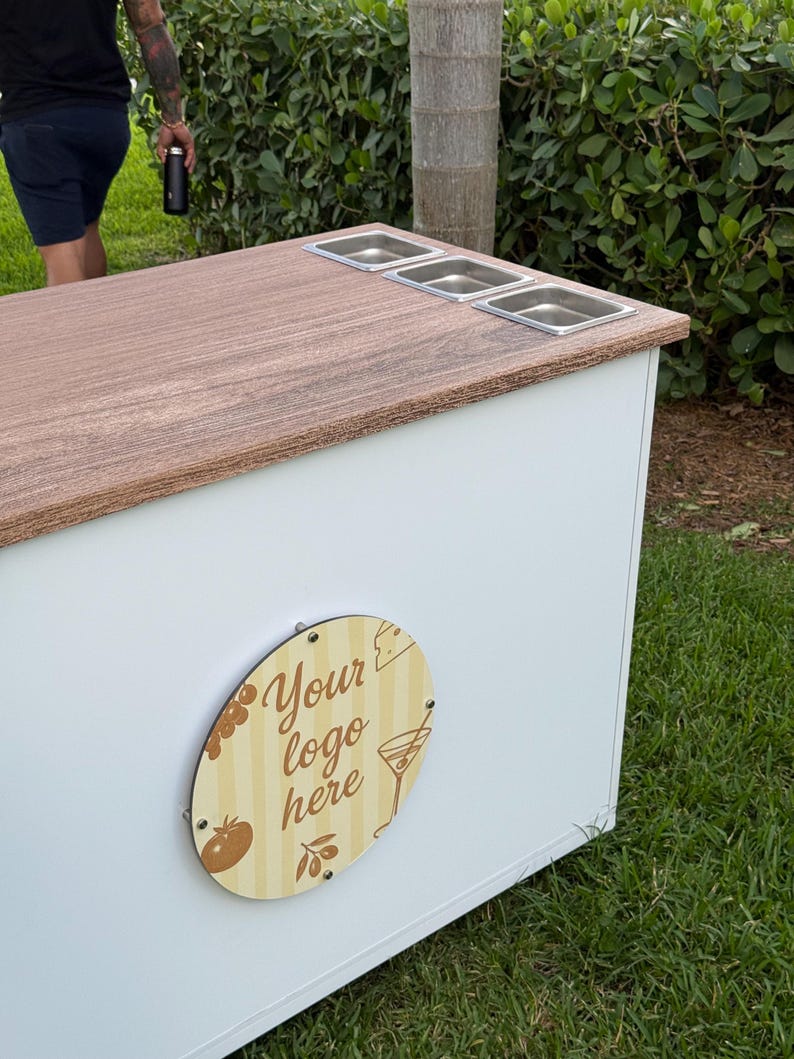 May include: A white and wood-toned outdoor bar with three stainless steel food containers. The bar features a wooden oval sign with the text "Your logo here" and food-related illustrations. The bar is set on a grassy area.