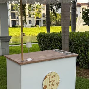 May include: A white outdoor bar with a wooden countertop and a white fringed umbrella. The bar has a white base and a wooden sign that says "Your logo here". The bar is set up on a grassy area.