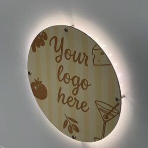 May include: A round, wooden sign with the text "Your logo here" in a script font. The sign features illustrations of olives, a tomato, cheese, and a martini glass. The sign is illuminated with a warm, white light.