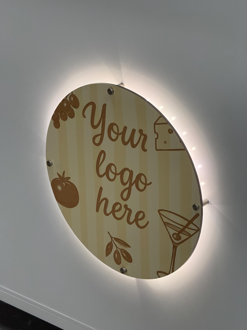 May include: A round, wooden sign with the text "Your logo here" in a script font. The sign features illustrations of olives, a tomato, cheese, and a martini glass. The sign is illuminated with a warm, white light.