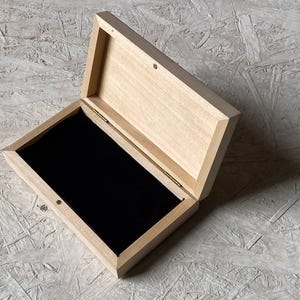 Pode incluir: Uma caixa de madeira clara aberta, revelando um interior de veludo preto. A caixa é retangular com uma tampa articulada. A madeira tem um grão natural e a caixa está sobre um fundo neutro. Ideal para guardar objetos.