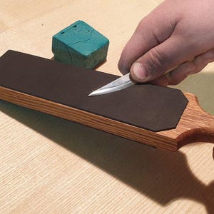 Puede incluir: Un asentador de madera con una superficie de cuero marrón oscuro se utiliza para afilar un cuchillo. Un pequeño bloque de compuesto de afilado verde está cerca. El cuchillo se desliza sobre la superficie de cuero.