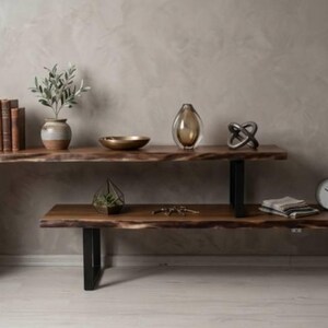 Pode incluir: Uma mesa consola de madeira de dois níveis com um design de borda viva. A prateleira superior contém itens decorativos como um vaso, uma tigela e uma escultura. A prateleira inferior apresenta um relógio e livros. A mesa é suportada por pernas de metal preto.
