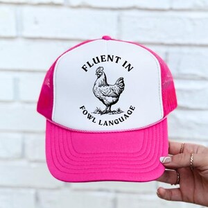 May include: A pink and white trucker hat with a black and white illustration of a chicken and the text "Fluent in Fowl Language".