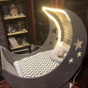 May include: A gray crescent moon-shaped crib with a white and gray chevron patterned mattress. The crib has a built-in light that illuminates the crescent moon shape. The crib is decorated with white stars.