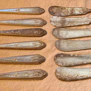 May include: A collection of antique butter knives, arranged in two rows. The knives have a silver finish with ornate handle designs. Some knives show signs of age and wear, with tarnishing and discoloration. The blades are wide and rounded.