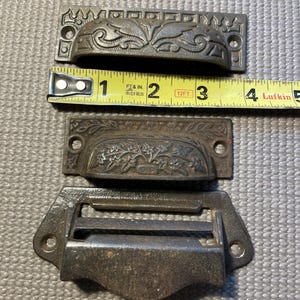 May include: Three antique metal drawer pulls in varying designs. The top pull is approximately 3 inches long, featuring an ornate floral pattern. The middle pull has a similar design, and the bottom pull is a simple, rectangular handle.