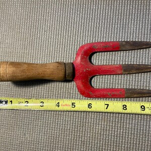 May include: A hand cultivator with a wooden handle and a red metal head featuring three tines. The tool is positioned above a yellow measuring tape, showing a total length of approximately 10 inches. The metal head is stamped with the text MADE IN ENGLAND.