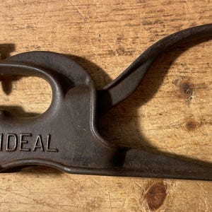 May include: A vintage, cast-iron tool with the word "IDEAL" embossed on its base. The tool features a curved handle, a pointed punch, and a screw for adjustment. The metal has a dark, aged patina, suggesting its use over time.