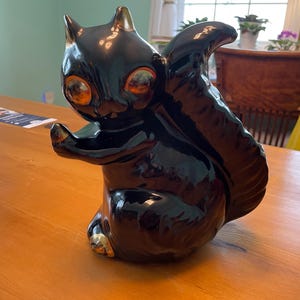 May include: A glossy black ceramic squirrel figurine with gold accents on its paws and eyes. The squirrel is in a seated position with its tail curved upwards. The figurine is on a wooden table.