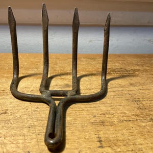 May include: A four-pronged metal hand cultivator featuring a dark, weathered, and rusted finish. The tool has a central handle loop and four upright, pointed tines designed for soil aeration and garden maintenance tasks.