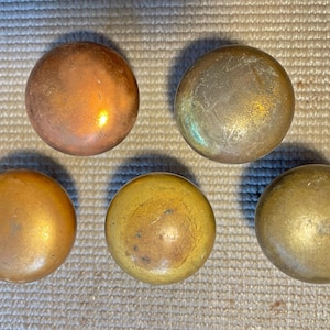 May include: Five antique brass knobs in varying shades of gold and copper. The knobs are round with a smooth, polished surface. The knobs are arranged on a textured, neutral-colored surface.