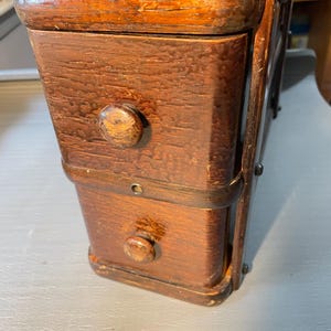 May include: A tall, narrow wooden storage unit with a rich brown finish, featuring two stacked drawers with small, rounded wooden knobs. The structure has a visible grain texture, rounded corners, and dark metal hardware accents along the sides.