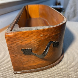 May include: A semi-circular wooden drawer with a smooth, polished finish and a prominent wood grain pattern. The front features a dark, ornate metal handle with decorative leaf-like ends, securely attached to the curved wooden surface.