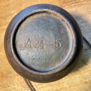 May include: A weathered, dark metal object with a rounded shape. The base features the stamped text "A24-5". The object has a dark, matte finish with visible signs of rust, suggesting age and use. A thin, dark cord is attached.