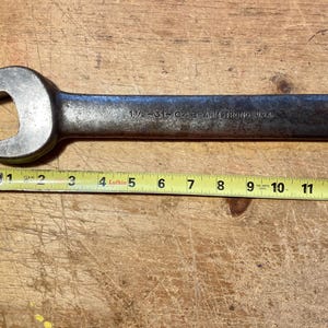 May include: A dark grey metal open-end wrench with the text 1 1/2-31-04B-ARMSTRONG U.S.A. stamped on the handle. A yellow measuring tape is positioned underneath the wrench, showing a total length of approximately 12.5 inches for the tool.