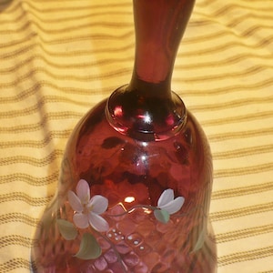 May include: A ruby red glass bell with a long handle. The bell is decorated with white and green floral accents. The bell is sitting on a striped fabric.