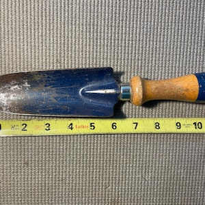 May include: A blue metal garden trowel with a wooden handle and a blue painted end cap. The trowel is positioned above a yellow Lufkin measuring tape, showing the tool measures approximately 10.5 inches in total length from tip to end.