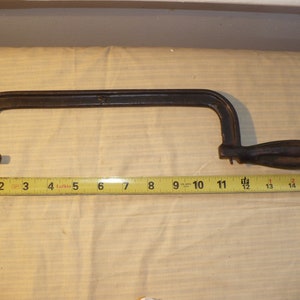 May include: A dark brown metal coping saw with a wooden handle and a brass wing nut, positioned above a yellow measuring tape that indicates a total length of approximately 15.5 inches from the handle to the end of the frame.