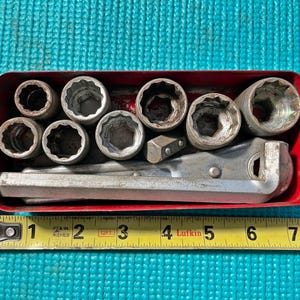 May include: A red metal toolbox containing a set of silver sockets and a ratchet wrench. The toolbox is open, revealing the tools inside. A yellow measuring tape is placed next to the toolbox. The toolbox is approximately 8 inches long.