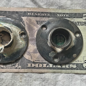 May include: Two black metal round decorative pieces with a white center and a hole in the middle. The pieces are sitting on a five dollar bill.