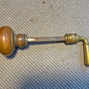 May include: An antique door handle with a copper-colored knob and a brass handle. The handle has a long, metal rod with a toothed edge. The handle is curved and has a cylindrical grip.