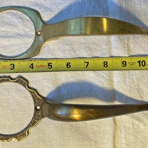 May include: Two vintage brass door handles. The top handle is approximately 11 inches long, and the bottom handle is approximately 12 inches long. Both have a circular grip and a decorative end. A yellow measuring tape is placed between the handles.