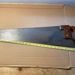 May include: A vintage hand saw with a wooden handle and a metal blade. The blade is approximately 27 inches long. A yellow measuring tape is placed along the saw's blade.