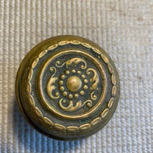 May include: Antique brass door knob with an ornate, circular design. The knob features a central medallion with a raised floral pattern and a beaded border. The brass has a weathered, aged appearance, suggesting vintage craftsmanship.