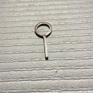 May include: A small, metallic, copper-toned key-like object featuring a circular ring at the top connected to a straight, narrow, rectangular stem. The piece has a visible split where the ring meets the stem, showing a simple, flat construction.