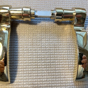 May include: A pair of shiny gold-colored door handles. The handles are connected by a metal rod. The handles have a curved design and reflect the surrounding environment. The door handles are likely for a cabinet or door.