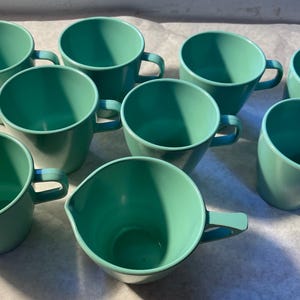 May include: A collection of eight teal-colored mugs with angled sides and curved handles. The mugs are arranged in a group, showcasing their uniform design and color. The mugs are made of a solid material.