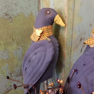 May include: A handcrafted, dark blue fabric bird with a yellow beak and a plaid collar. The bird has a decorative leaf accent on the collar. The bird is part of a fall-themed decorative display.