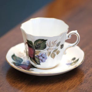 May include: A white teacup and saucer set with a floral and fruit pattern. The cup and saucer have a gold trim. The design features green leaves, purple grapes, and red berries. The teacup has a distinctive, scalloped edge.