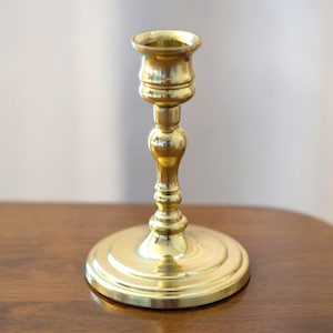 May include: A polished brass candlestick holder featuring a wide, circular stepped base, a slender central stem with decorative rounded nodes, and a flared cup at the top designed to securely hold a single taper candle in an upright position.