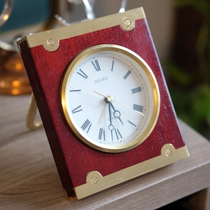 May include: A rectangular Seiko clock with a red wooden frame and gold-coloured accents. The clock face is white with black Roman numerals and gold hands. The clock is resting on a wooden surface.