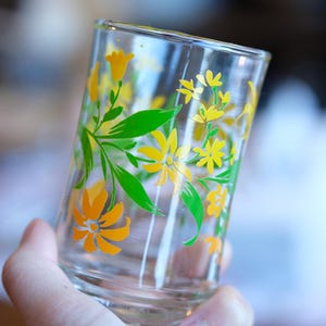 Può includere: Un bicchiere di vetro trasparente decorato con disegni floreali gialli e arancioni e foglie verdi. Il bicchiere è tenuto in mano ed è leggermente angolato, mostrando il motivo floreale. Il bicchiere viene probabilmente utilizzato per bere.